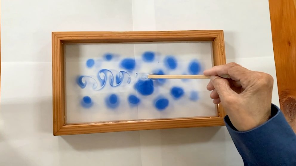 Jennifer Greene drawing a slender stick through blue dye in a shallow tray during the Train of Vortices experiment